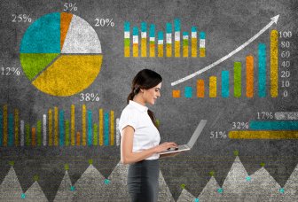 Photo of a woman standing in front of some graphs and tables, holding a laptop.