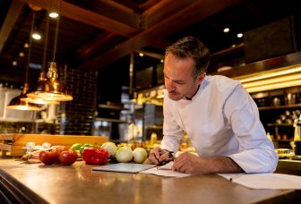 Chef looks at iPad while jotting down on a notepad. Photo.